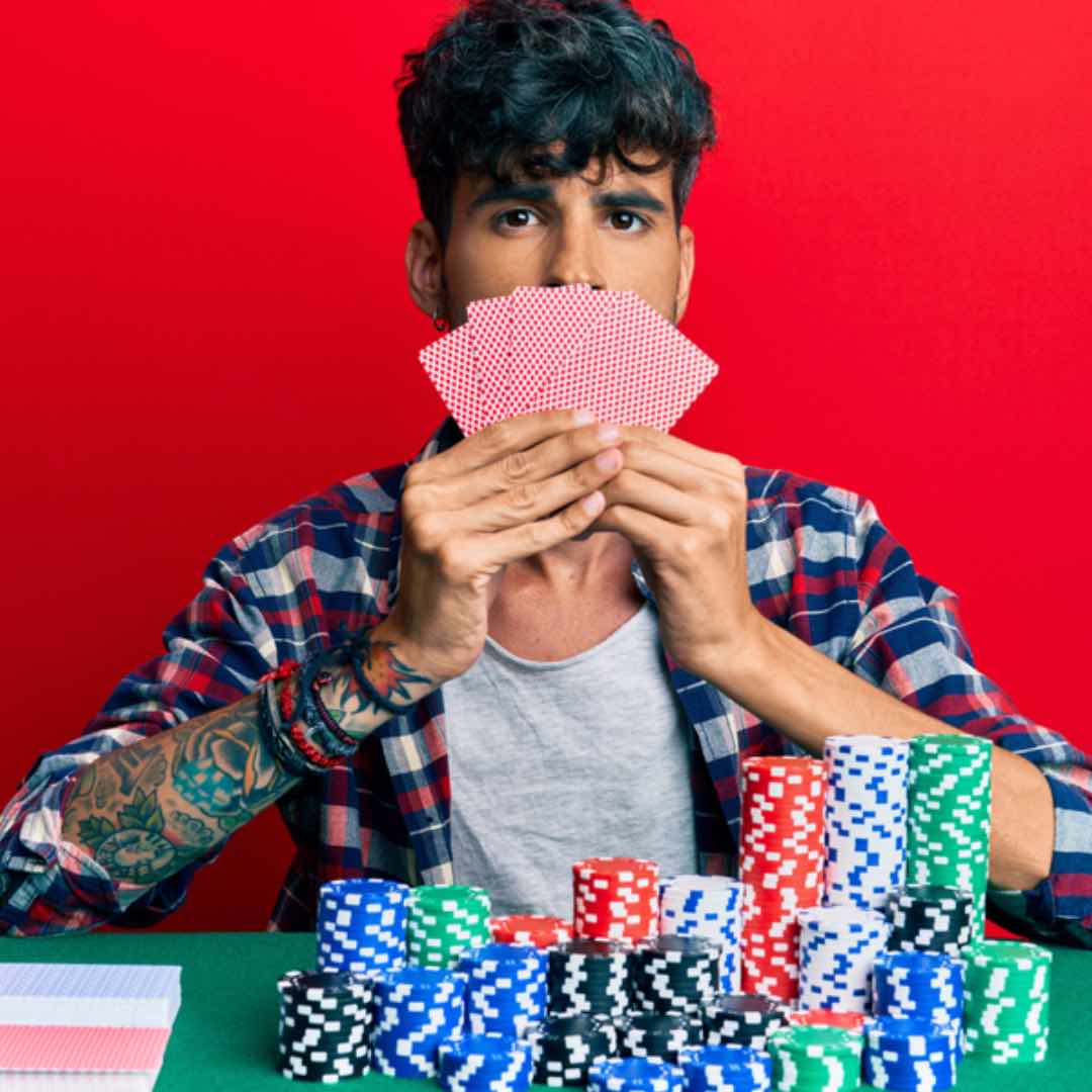 A poker player in front of a red wall and piles of poker chips holds his cards over his mouth.