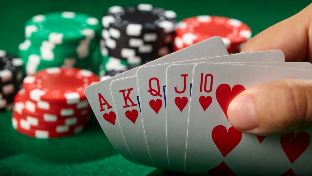 A person holding up a royal flush in hearts in front of some chips on a poker table.