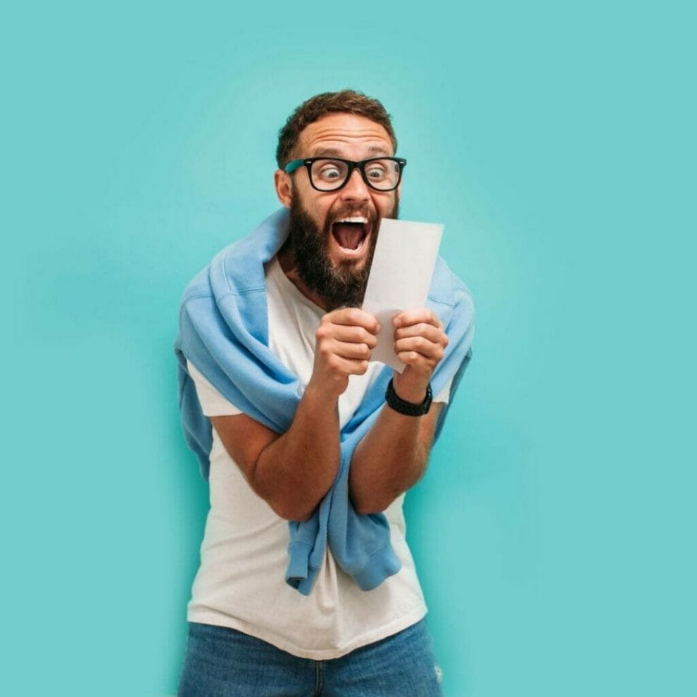 A man against a blue background celebrating while looking at a lottery ticket.