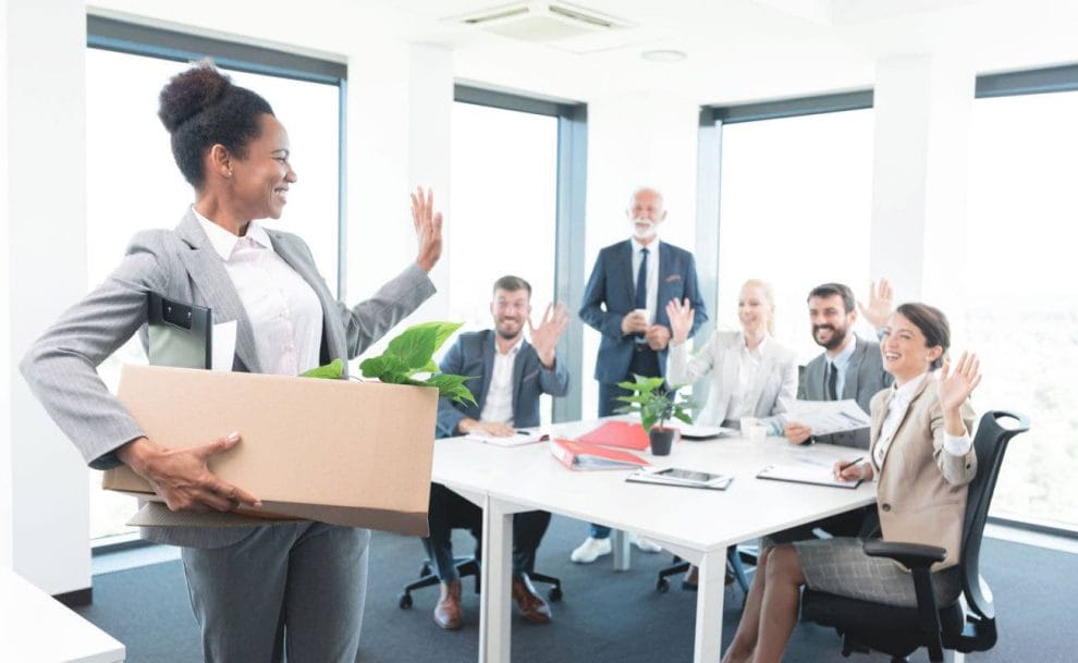 A woman in a suit holding a box and waving goodbye to her colleagues in the boardroom.