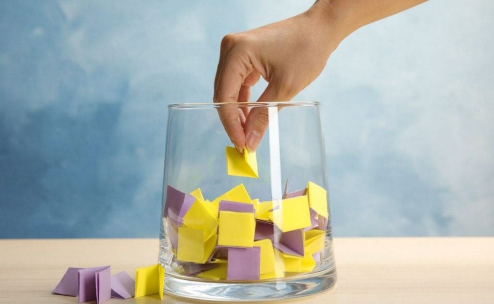 A hand takes out a folded yellow paper from among other folded yellow and purple papers in a glass vase.