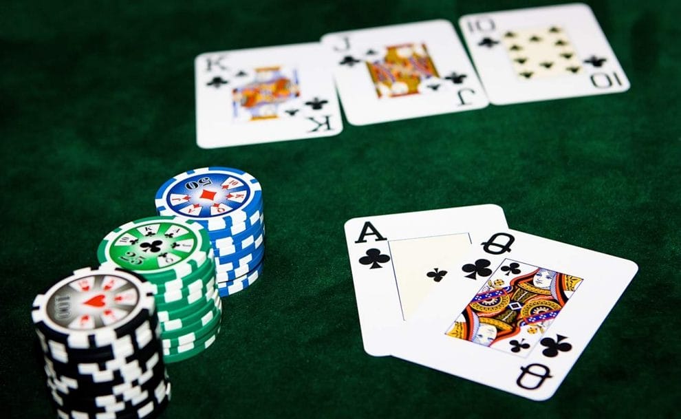 Two hole cards are revealed next to several stacks of poker chips. The two hole cards are an ace and a queen of clubs. In the background the community cards are blurry but visible. They are a king, jack and 10 of clubs.