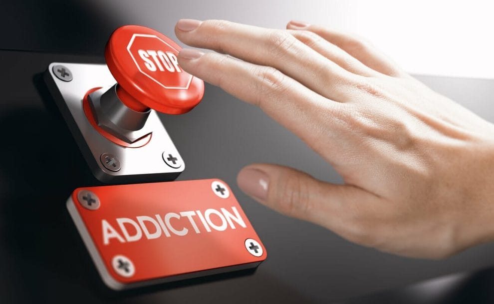 A hand reaching for a red stop button with a sign reading ‘addiction’ under it.