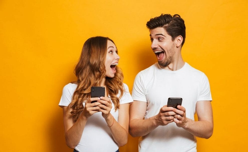 A man and woman holding smartphones look at each other with elated expressions.