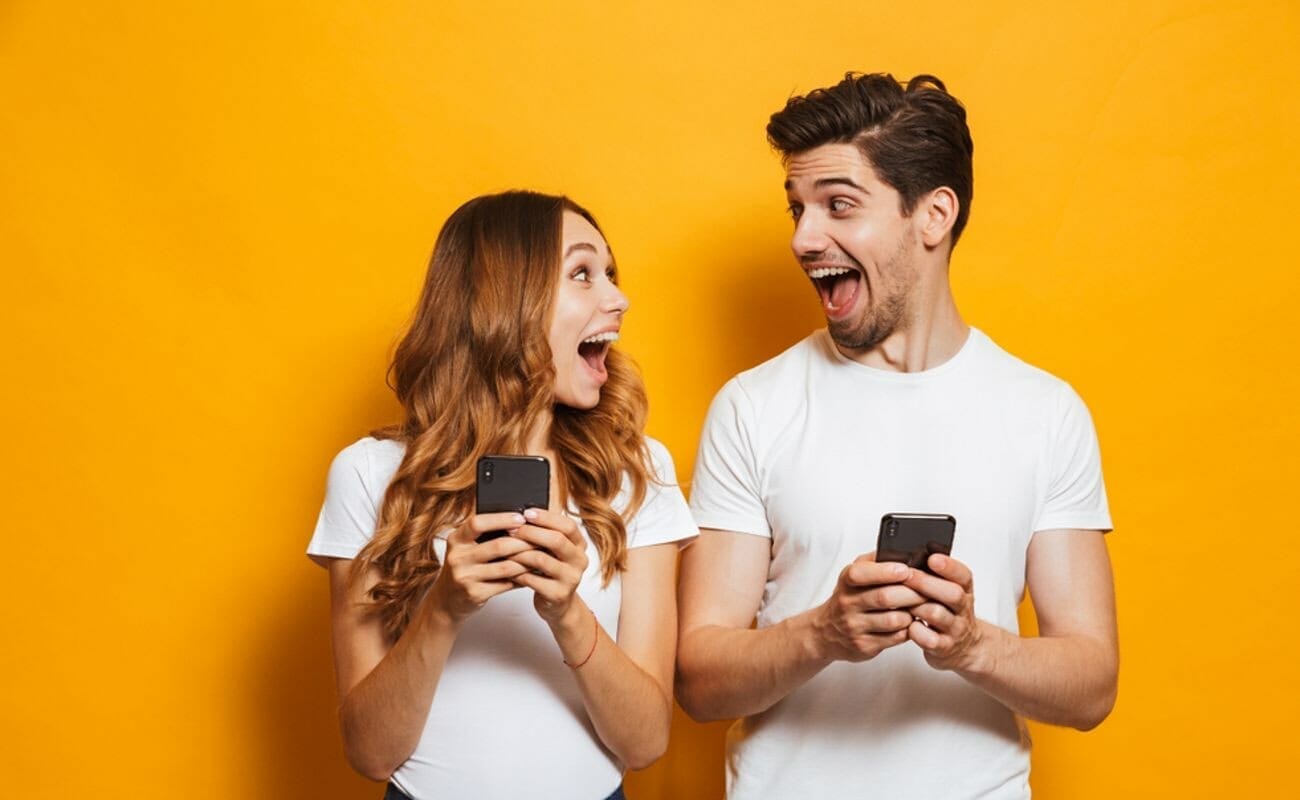 A man and woman holding smartphones look at each other with elated expressions.