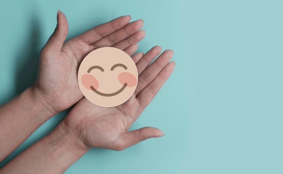 A hand holding a nude color paper cutout smile on a turquoise background.
