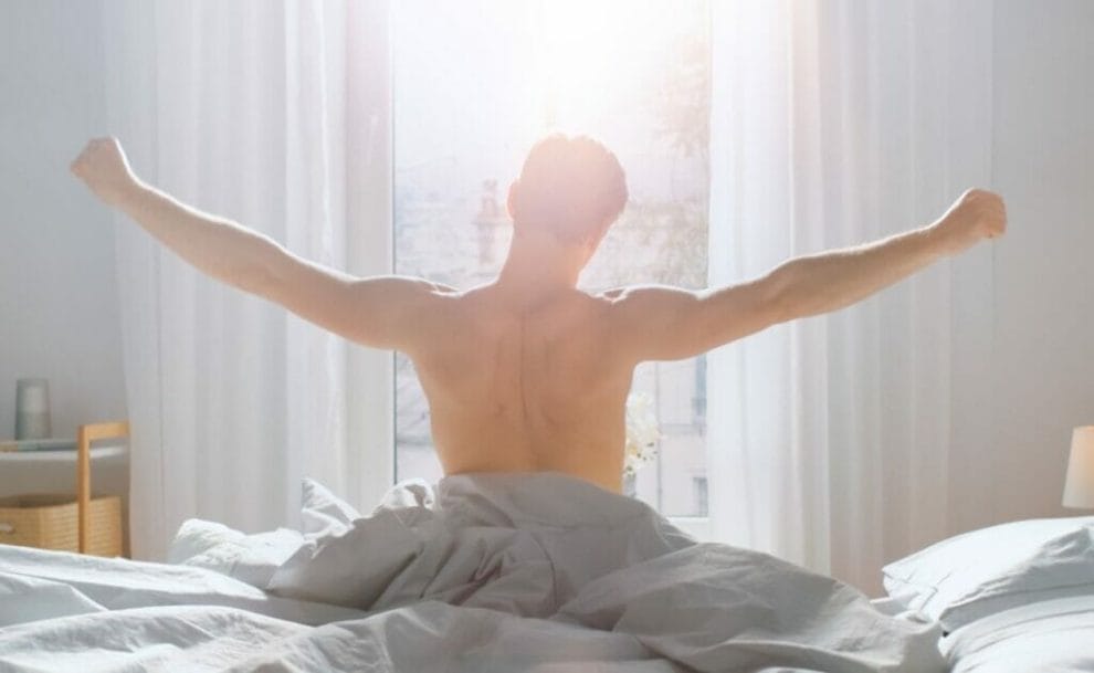 A man getting out of bed stretching in front of a large sunny window.