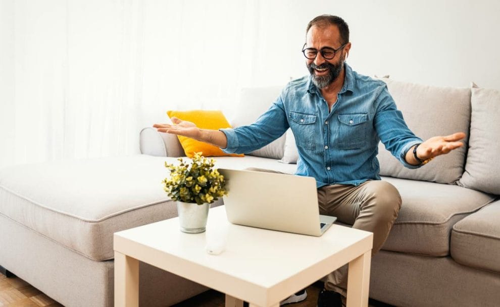 A happy person relaxes on the couch with a laptop on the table.