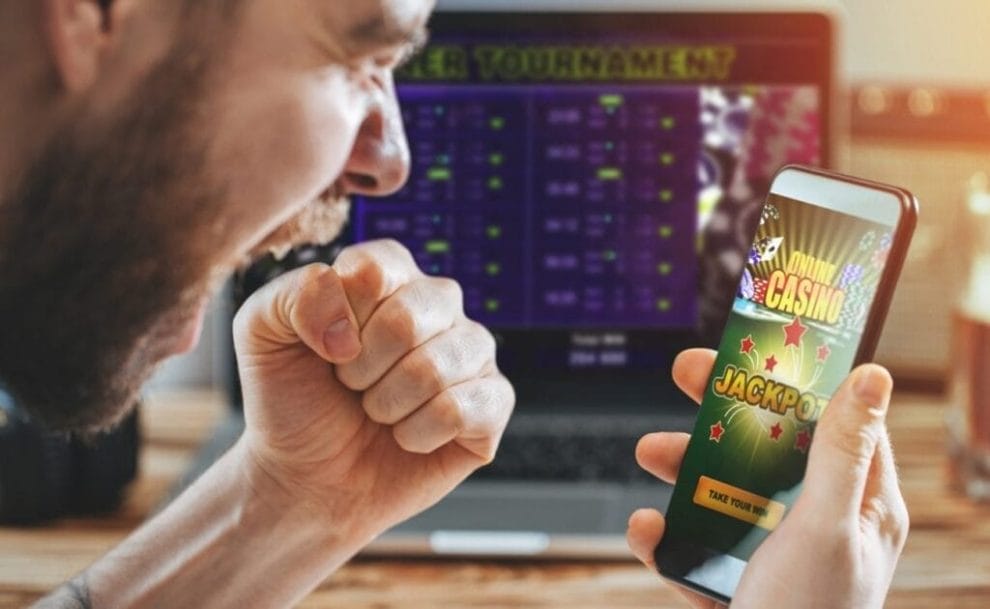 A man celebrates winning an online jackpot on his phone, a laptop on an online casino in the background