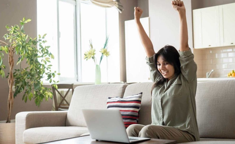 A woman celebrating with her hands in the air at her laptop on the couch.