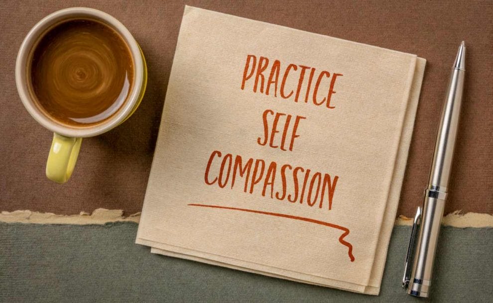 A coffee, a page with the words “practice self-compassion” and a pen on a table.