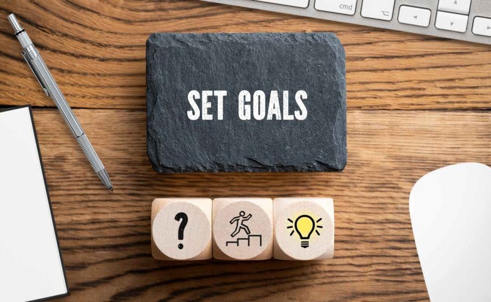 A black stone with the words “Set Goals” on a wooden table with a pen and keyboard.