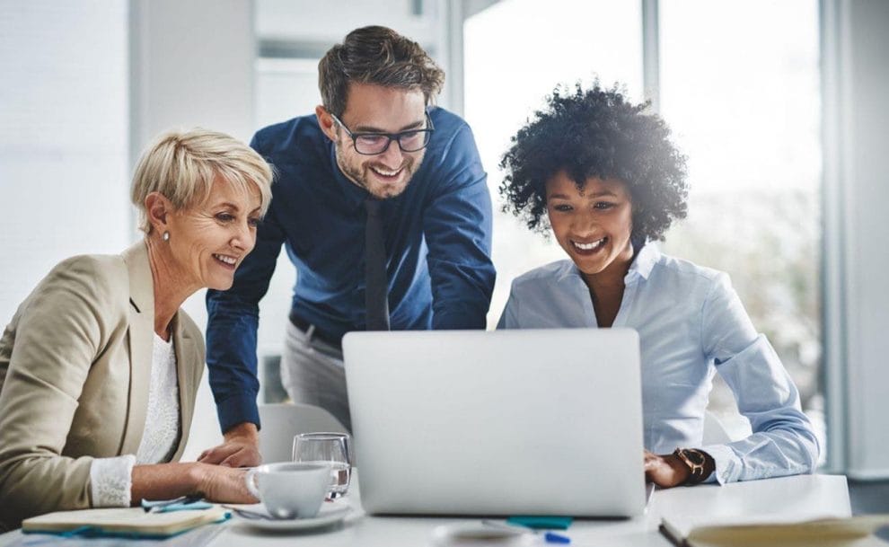 Three coworkers smile while looking at a laptop screen.