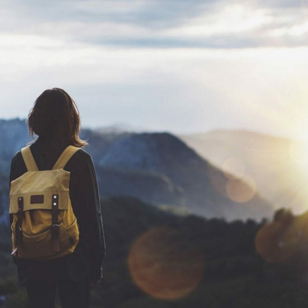 A traveler wearing a backpack overlooks a mountain range with the sunrise in the background.