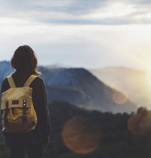 A traveler wearing a backpack overlooks a mountain range with the sunrise in the background.