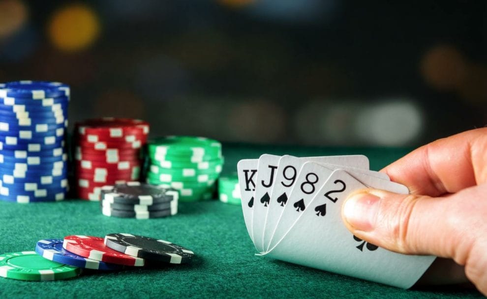 Next to a stack of poker chips, a hand reveals a hand of playing cards over the green surface of a poker table.