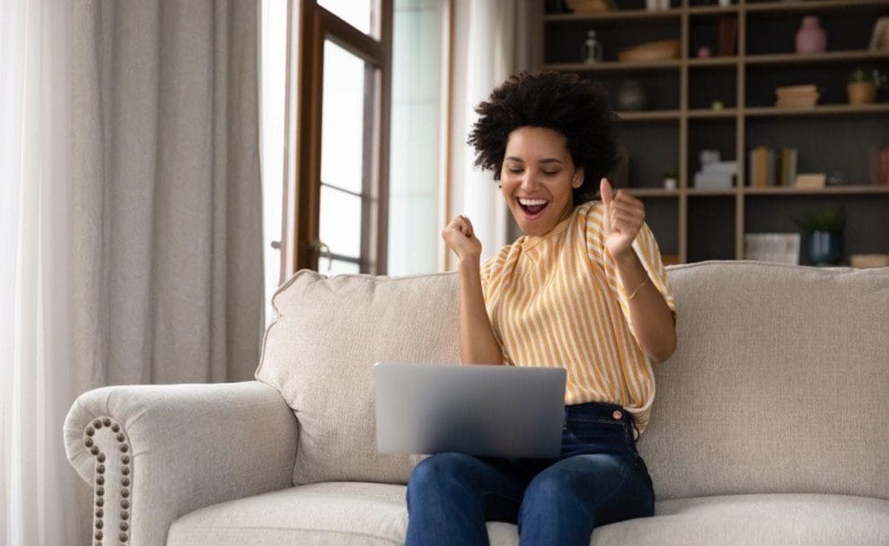 A woman celebrating a win while sitting on her couch looking at a laptop screen.
