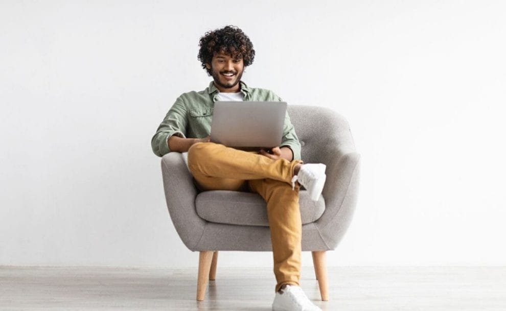 A cheerful person sits on a lounge chair looking at their laptop screen.