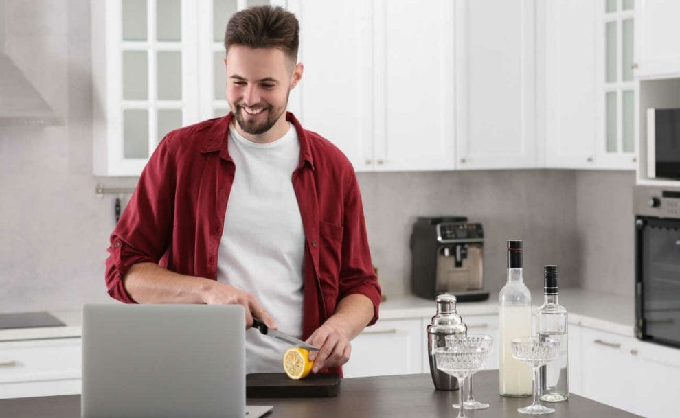 A person smiles at their laptop while making cocktails in their kitchen.
