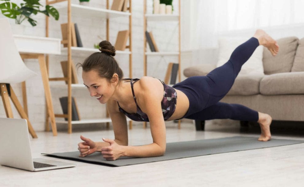 A person looks happily at their laptop while performing yoga in their lounge.