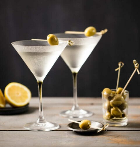 Two martinis on a wooden table next to lemons and a glass of olives.