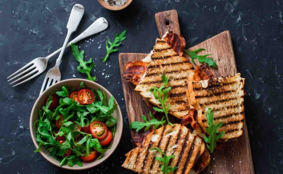 A board with three grilled sandwiches on it sits next to two forks and a bowl of salad.