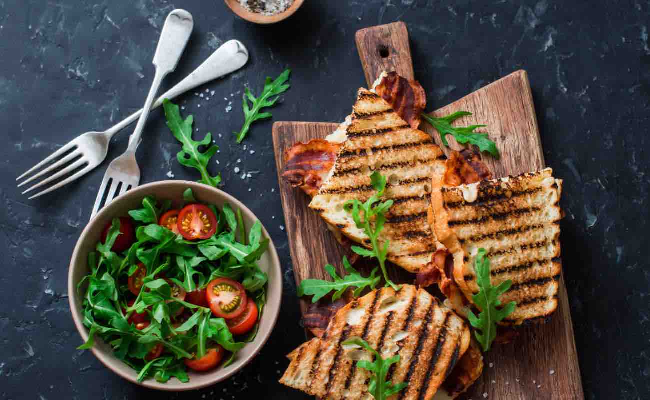 A board with three grilled sandwiches on it sits next to two forks and a bowl of salad.