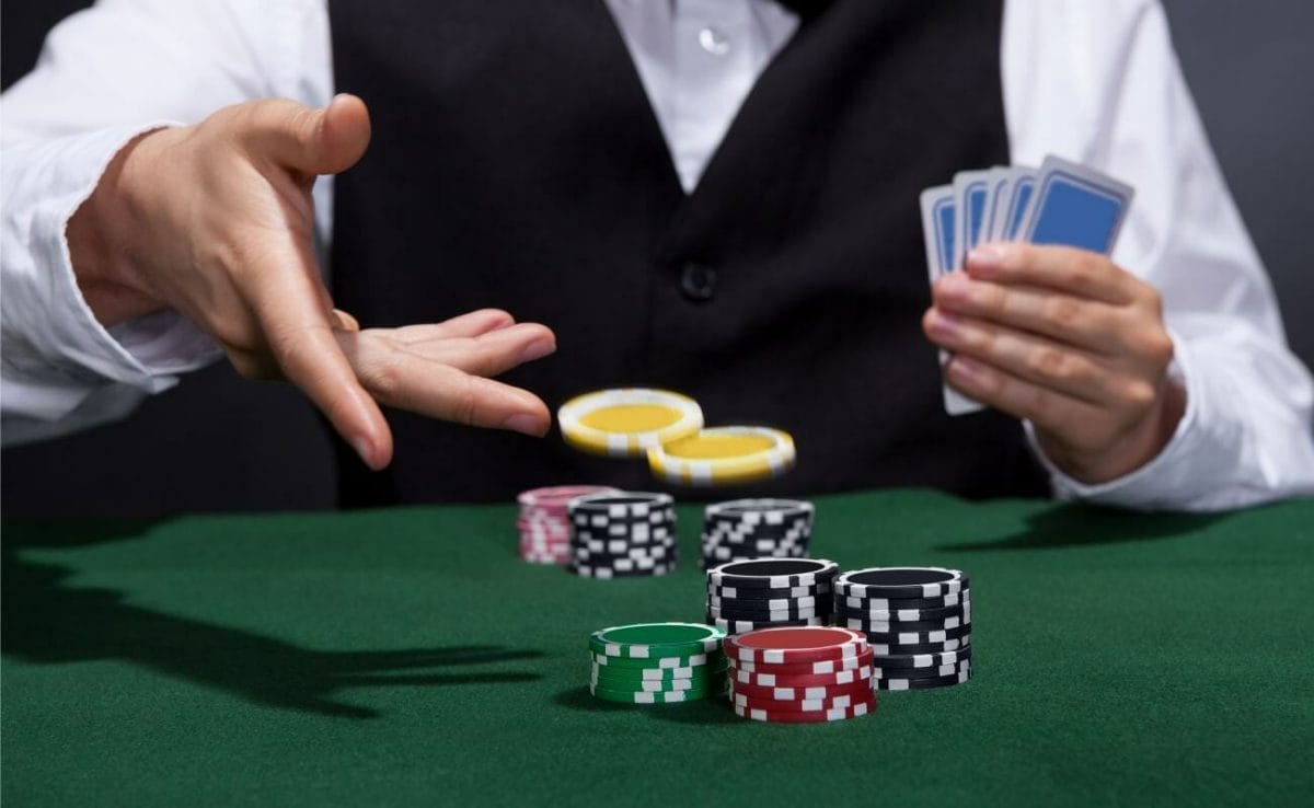 A person wearing a black waistcoat over a white shirt throws casino chips while holding playing cards.