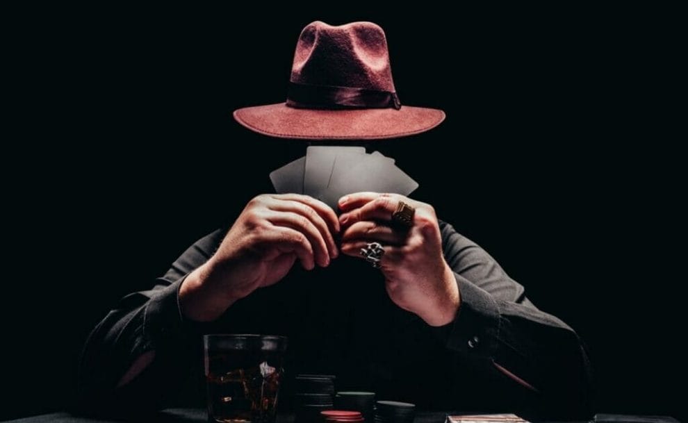 A mysterious man wearing a hat holding playing cards to his face at a poker table.
