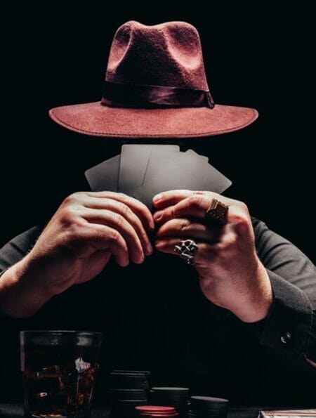 A mysterious man wearing a hat holding playing cards to his face at a poker table.