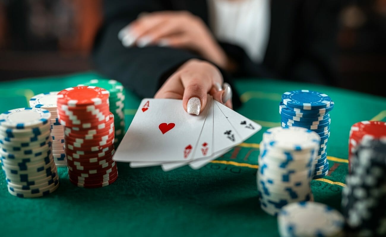 A person with long, shiny nails reveals their poker hand of four aces. There are stacks of poker chips on either side of the cards.
