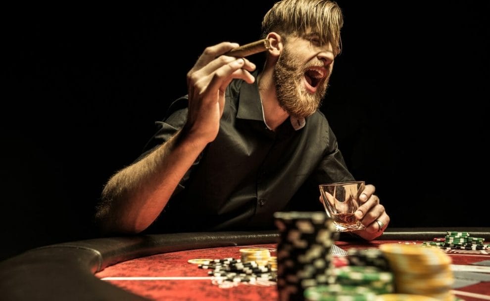 A person speaks loudly while holding a cigar and a drink at a casino game table.
