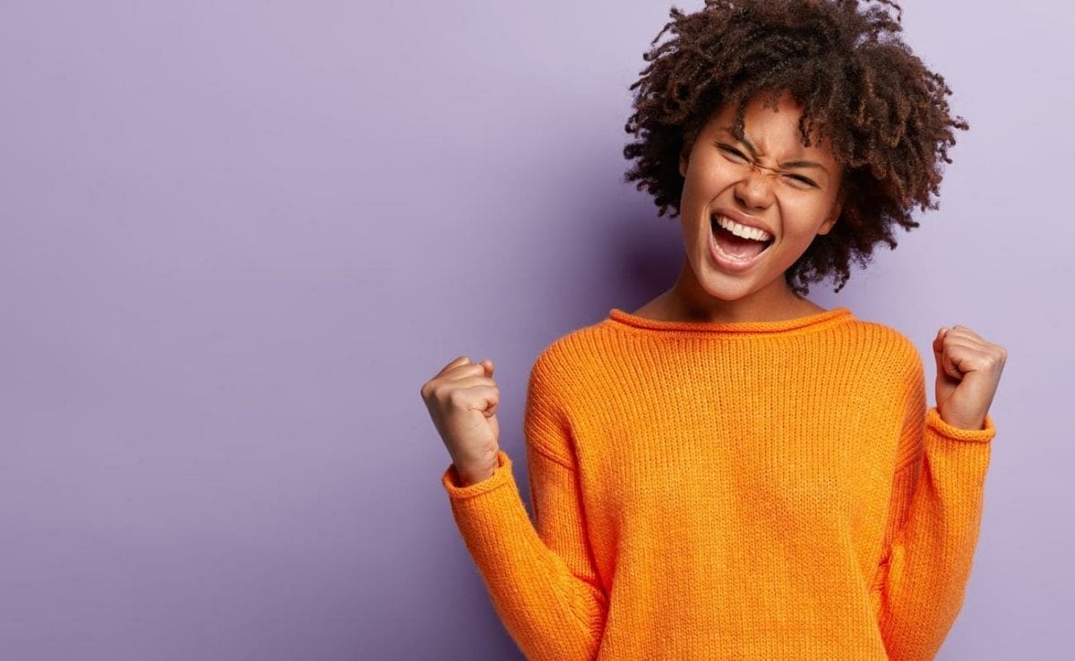 A person wearing an orange knit sweater cheers with their fists raised.