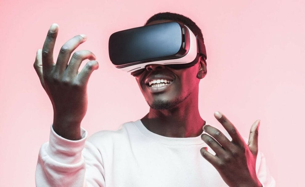 A person wearing VR goggles and a white crewneck T-shirt raises their hands in front of them.
