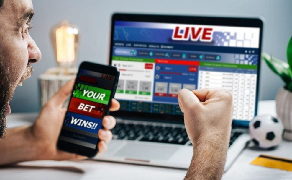 Man celebrating his online sports betting win, he is holding a mobile device in his hand that states “your bet wins” while he cheers at the laptop screen displaying the live sport betting tournament 
