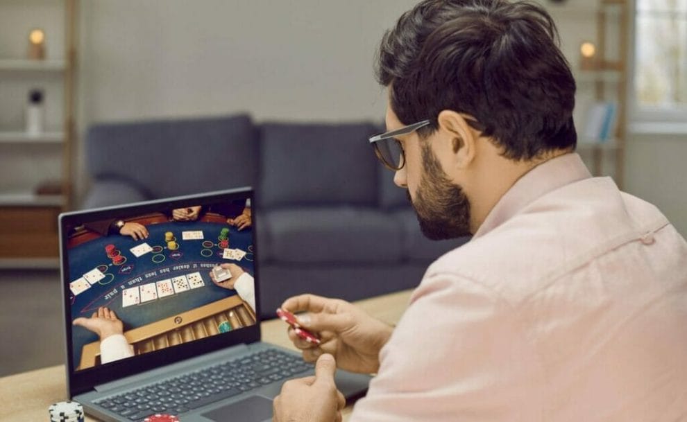 A man playing online Blackjack on a laptop at a desk holding poker chips in his right hand with more poker chips stacked next to the laptop