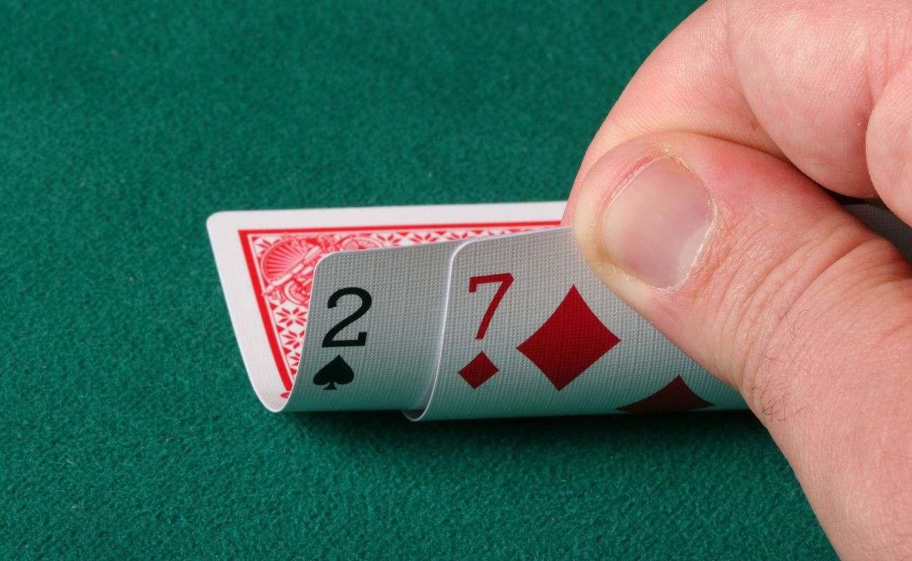 a close up of a man’s hand checking his hole cards, a spade two and diamond seven, during a game of poker on a green felt poker table 