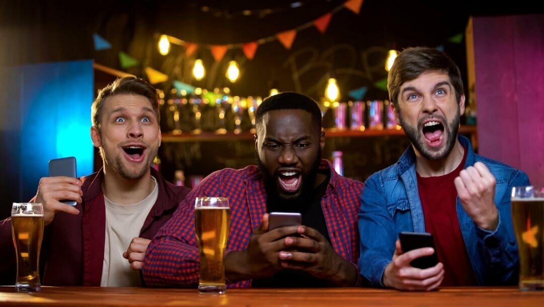 three excited males gripping their phones while  cheering for their sports team at a bar with a beer in front of each man