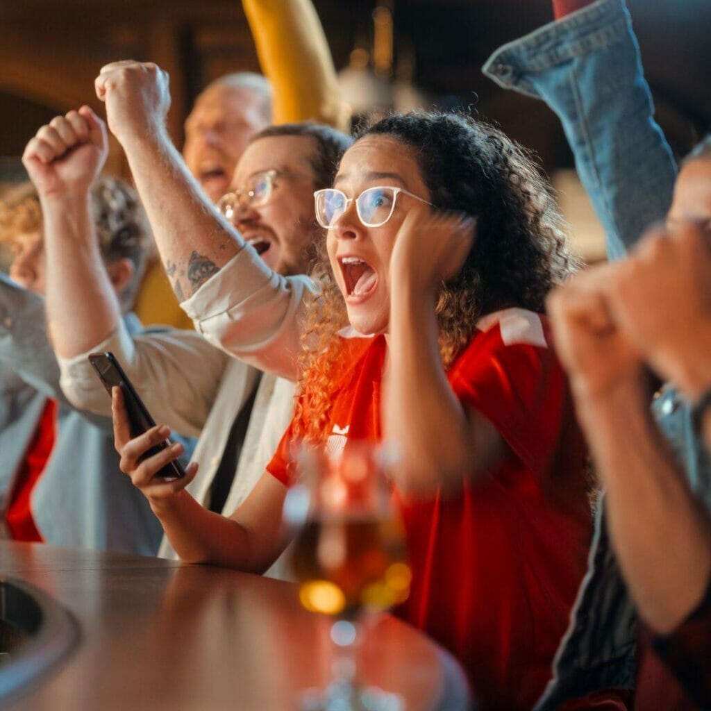 a woman holding a phone surrounded by men, all are cheering for a sports team at a bar