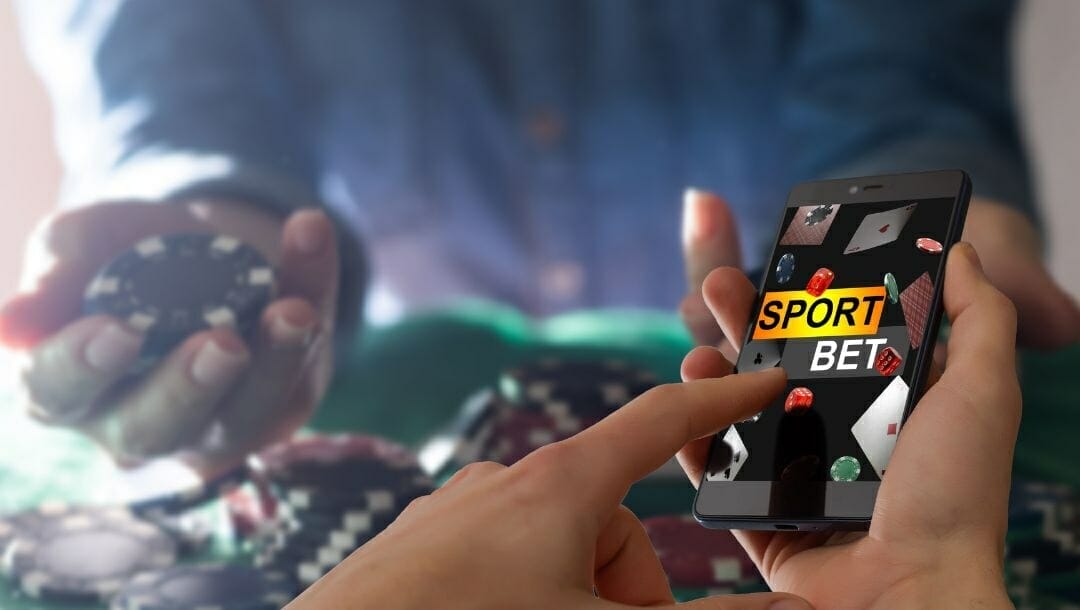 a person holding a phone making a sport bet online while the person in the background is holding stacks of poker chips