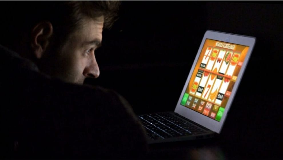a man in a dark room, resting his head on his arms on a table while staring at a laptop in front of him that displays an online slots game