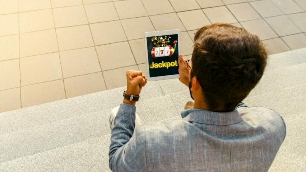 the back view of a man sitting on steps as he celebrates his jackpot win displayed on the digital tablet that he is holding with his right hand, he is playing online slots 
