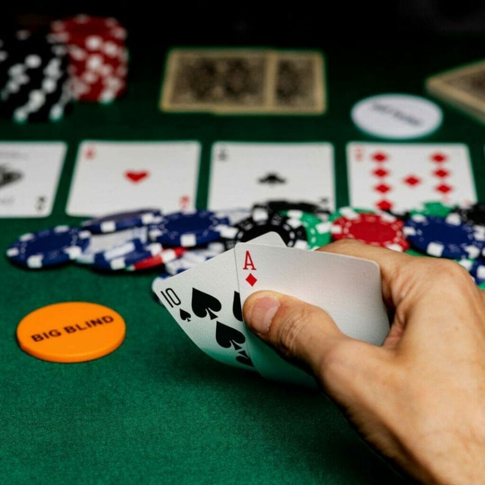 Header image, close up of a man’s hand checking his hole cards with four cards turned over in front of him and poker chips scattered around, a bright orange “BIG BLIND” token in front of him