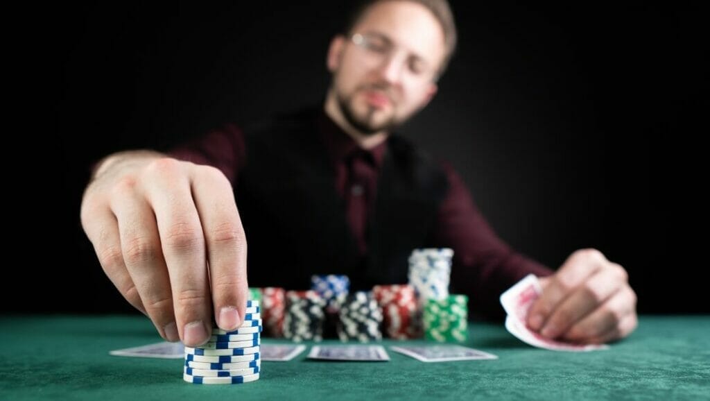 A man sitting at a poker table with poker chips stacked and four playing cards fanned face down in front of him, his left hand flips up two hole cards and his right hand fixes a stack of white and blue poker chips