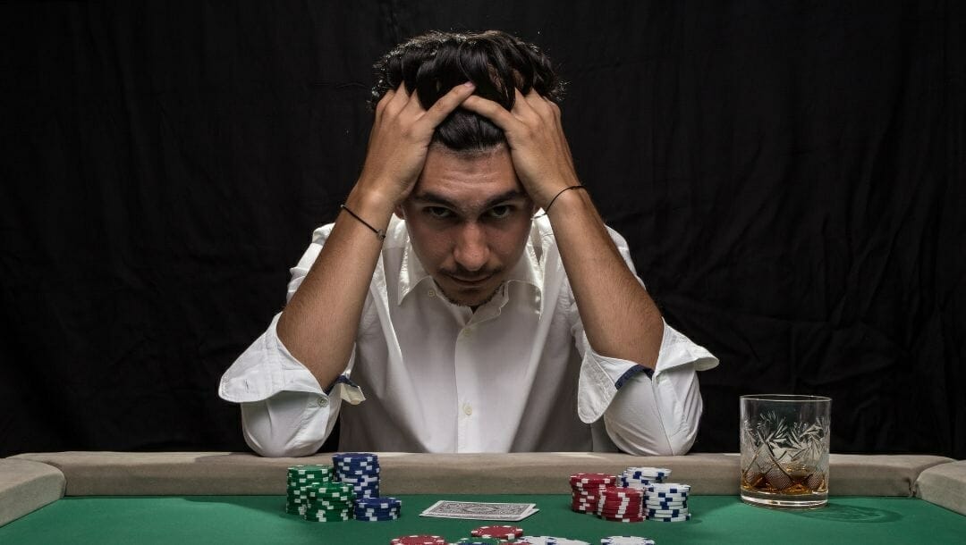 Body image, a man sitting at a poker table with his head in his hands and a stressed expression on his face, poker chips and cards in front of him on the table, a drink beside him 