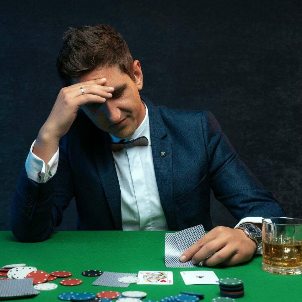 Header image, man sitting at a green poker table looking at his hole cards with his left hand, his right hand in on his head in a stressed expression, a drink to his left, cards and poker chips are scattered in front of him