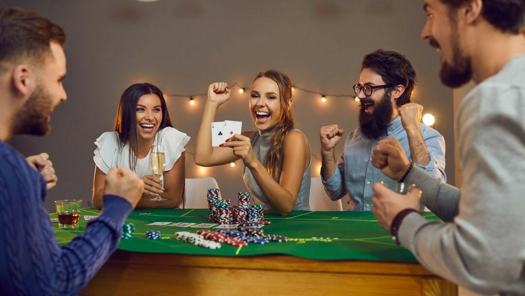 Body image, five friends celebrating a poker win as they sit around the poker table cheering, poker chips stacked in the middle of the table, string lights on the wall behind them 