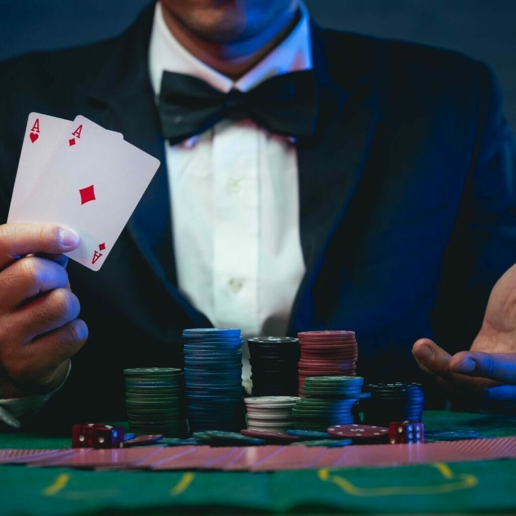 Header image, man sitting at a poker table holding a pair of aces, poker chips stacked and playing cards fanned out face down in front of him