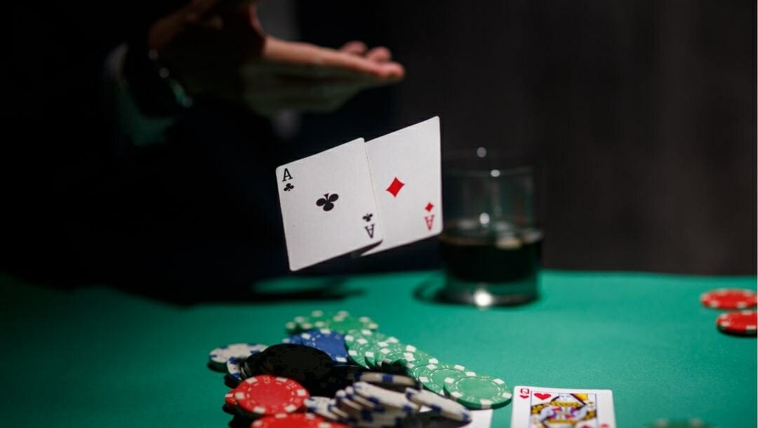 Body image, poker chips and cards scattered on a poker table, a man’s hand throwng a pair of aces, a drink in the background 
