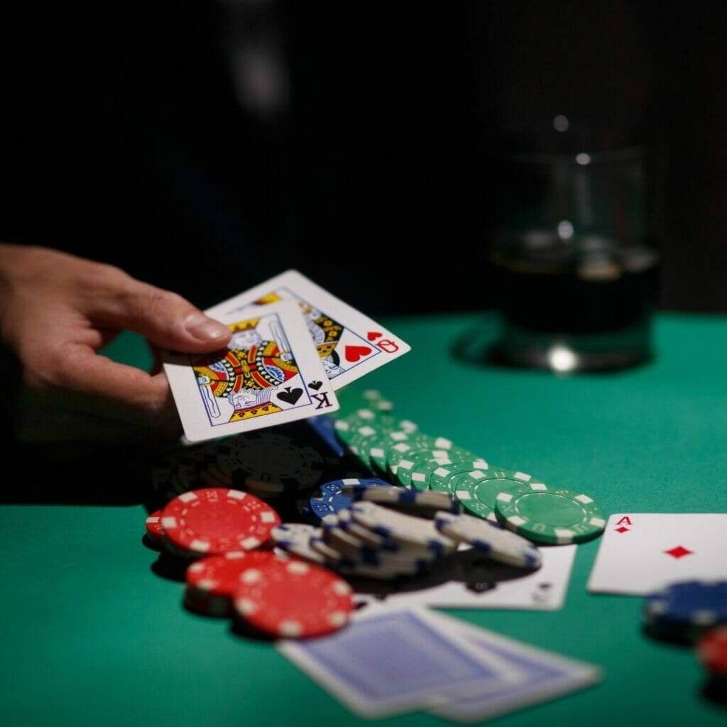 Header image, poker chips and cards scattered on a poker table, a man’s hand holding out a king of spades and queen of hearts, a drink in the background